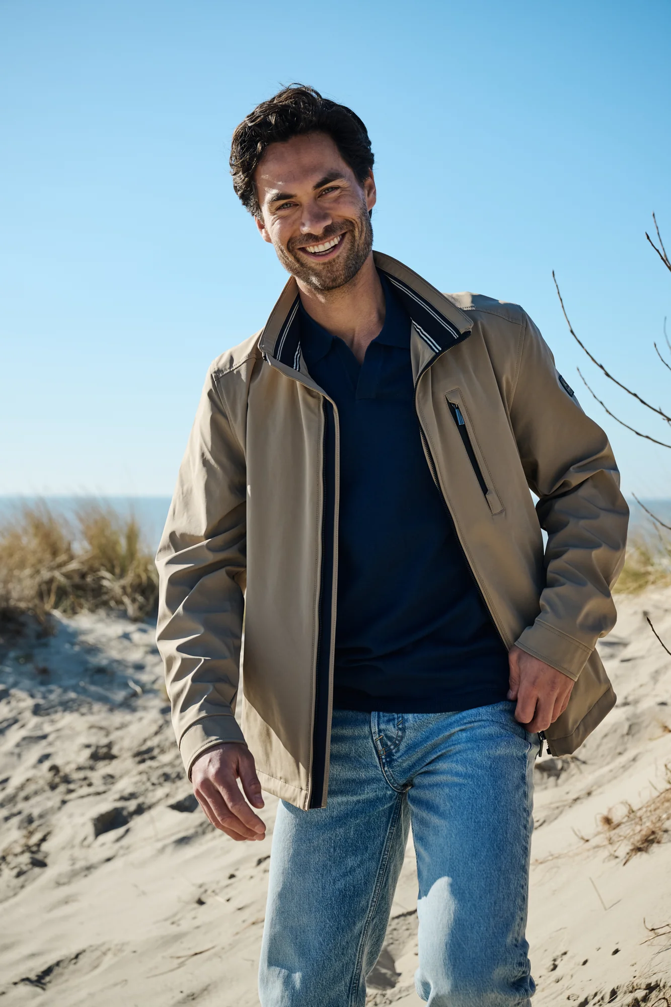 Raphael Korse lachend in een beige jas tussen de duinen